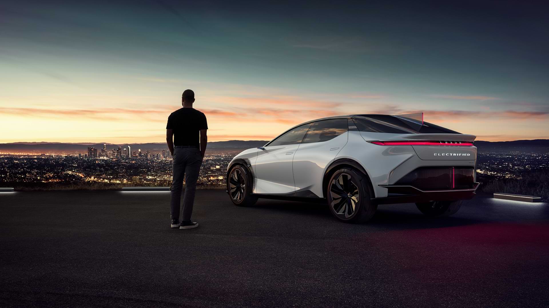 Man stands overlooking lit up city, with the LF-Z concept vehicle to his right.