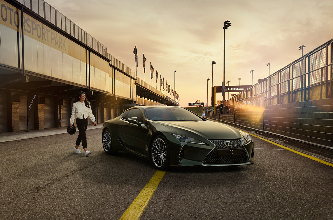 A woman dressed in a white jacket and black pants and holding a helmet walks towards an LC coupe parked in a pit lane