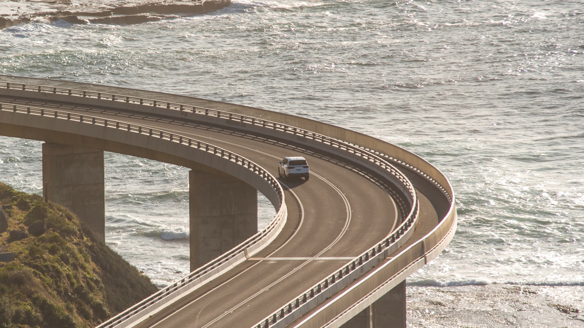 Wide, rear shot of a white Lexus LX driving across a beach-side bridge.